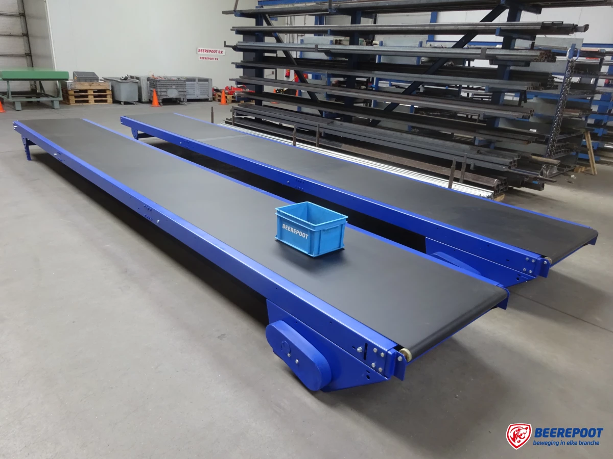 Je ziet twee blauwe vlakke bandtransporteurs van Beerepoot netjes naast elkaar in een voorraadhal van Beerepoot. Op een bandtransporteur staat een blauw Beerepoot kratje. Op de achtergrond zijn diverse onderdelen van machines in beeld.